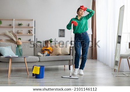 Tired mother taking break while cleaning at home, woman leaning on mop and touching head, kid son sitting on sofa and watching cartoon on smartphone. Lady exhausted of tidying up house