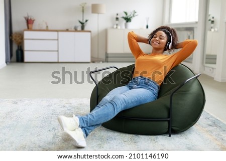 Rest And Relax Concept. Calm black woman sitting on bean bag, listening to music, audio book, podcast, enjoying meditation for sleep and peaceful mind in wireless headphones, leaning back, copy space Royalty-Free Stock Photo #2101146190