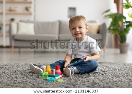 Happy little toddler boy watching cartoon on smartphone while sitting on floor carpet, looking and smiling at camera, playing with wooden colorful stacking toys, free space