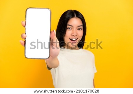Lovely excited chinese positive brunette young woman in casual t-shirt showing modern smart phone with blank white screen, standing on isolated yellow background, smiling. Mockup, copy space Royalty-Free Stock Photo #2101170028