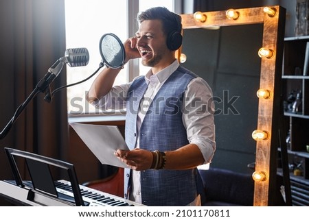 Young handsome bearded man is laughing at a home music studio in front of a piano with a makeup mirror behind his back.