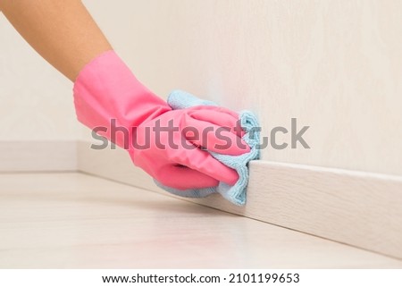 Young adult woman hand in pink rubber protective glove using blue dry rag and wiping light wooden baseboard surface in room at home. Closeup. Royalty-Free Stock Photo #2101199653
