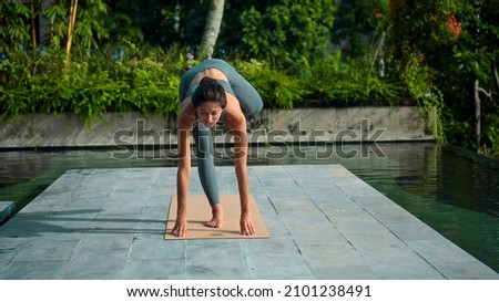 A young attractive European brunette sports girl makes yoga in nature with a green rainforest on a lit background by the pool on a mat and breathes deeply.