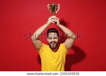 Excited jubilant exultant smiling fun young bearded man football fan in yellow t-shirt cheer up support favorite team hold champion cup under head isolated on plain dark red background studio portrait