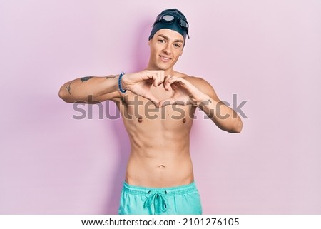 Young hispanic man wearing swimwear and swimmer glasses smiling in love doing heart symbol shape with hands. romantic concept. 