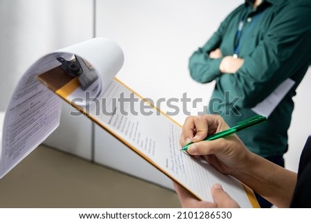 Inspector writting on a clipboard. Man in the background with arms crossed. Inspection by supervisors in a office Royalty-Free Stock Photo #2101286530