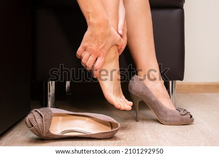 Pain in feet from uncomfortable shoes. A woman holds a sore foot with hands sitting on a couch. Ache caused by wearing high-heeled shoes. Discomfort from walking in heels for a long time. Orthodontics Royalty-Free Stock Photo #2101292950
