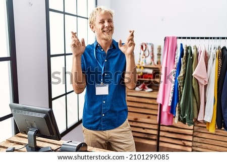 Young blond man working as manager at retail boutique gesturing finger crossed smiling with hope and eyes closed. luck and superstitious concept. 