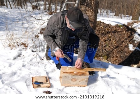 A man with a hammer in his hands sits and crafts a birdhouse against the background of a snowy forest. Nature preservation. Ecology concept.