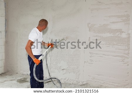 A man is applying stucco on the wall, plastering, coating the wall by spraying stucco with a stucco sprayer. Royalty-Free Stock Photo #2101321516