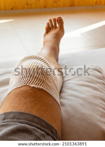VERTICAL, POV: Looking down at your knee wrapped in white bandages after a meniscus surgery. Male patient in home care after knee drainage surgery rests his leg on his leather couch. Post-surgery rest Royalty-Free Stock Photo #2101363891