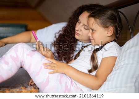 Smiling young girl and mother holding phone in left hand lying on bed both looking at phone in wooden house wearing pajamas in daytime. Family time together.