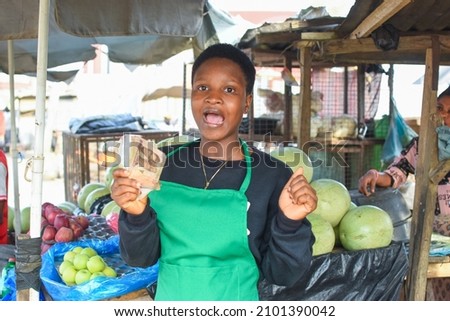 A happy African woman or female trader in green apron, holding some Naira notes or cash while standing at her stall in a local market