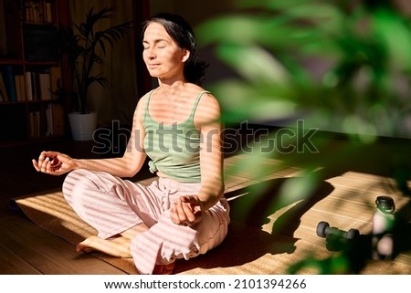 Woman practicing yoga and meditation at home sitting in lotus pose on yoga mat, relaxed with closed eyes. Mindful meditation concept. Wellbeing. Defocused foreground. Royalty-Free Stock Photo #2101394266