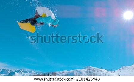 LENS FLARE: Stunning action shot of a snowboarding pro doing a spinning grab trick while riding in the sunny Julian Alps. Male snowboarder jumps high in the air and does a stunning rotating stunt.