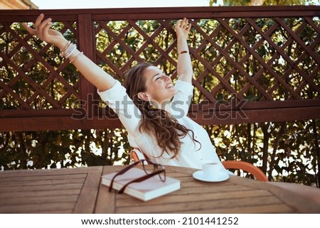 happy modern woman in white shirt with cup of coffee, book and eyeglasses sitting at the table in the patio of guest house hotel with raised arms rejoicing. Royalty-Free Stock Photo #2101441252