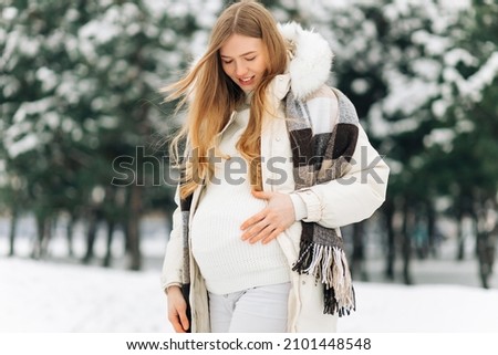 pregnant woman in warm winter clothes standing outside on a snowy winter day, with hands on her stomach on the background of nature. Concept of pregnancy, motherhood, expecting the birth of a child