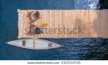 Aerial view of two Adirondack chairs on a wooden dock facing the blue water of a lake in Ontario on a sunny summer morning. A yellow canoe is tied to the dock. Life jacket and oars are visible. Royalty-Free Stock Photo #2101514725