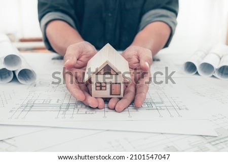 Architect engineer hands hold and protect mini house model on bueprint. Protect House, cost planning design and construction concept.