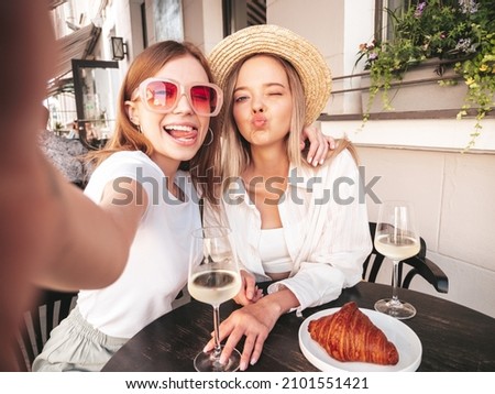 Two young beautiful smiling hipster female in trendy summer clothes.Carefree women posing at veranda cafe in the street.Positive models drinking white wine.Eating croissant.Taking Pov selfie photos Royalty-Free Stock Photo #2101551421