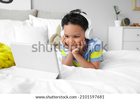 Little African-American boy with headphones watching cartoons on tablet computer in bedroom