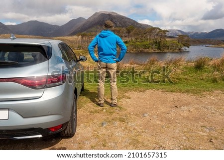 Male tourist standing by a car looking at a beautiful scenery. Twelve pine island, county Galway, Ireland. Mountains in the background. Travel and holiday concept. Cloudy sky. Copy space.