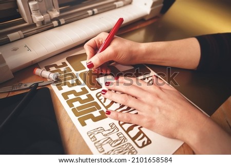woman with red painted nails removes excess material with weeding tool to make 2022 tiger design stickers from gold vinyl foil. workplace with cutting machine, tools, tablet computer. selective focus Royalty-Free Stock Photo #2101658584