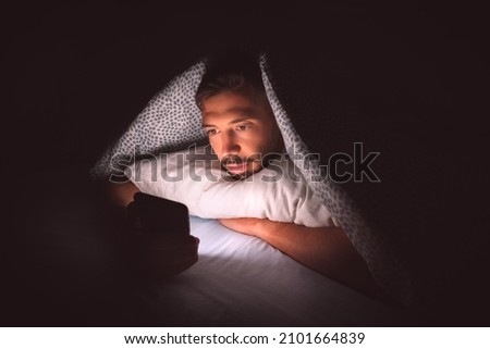 Young man lying in bed under a blanket and using smartphone at night. Social media addiction and communication Royalty-Free Stock Photo #2101664839