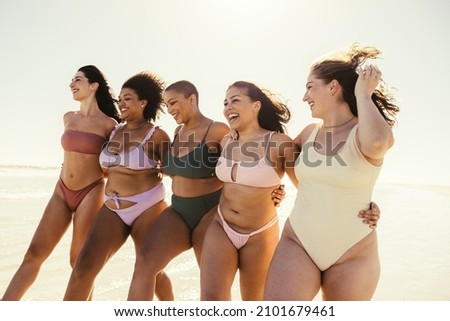 Summer fun in our bikini bodies. Happy young women embracing each other while wearing swimwear at the beach. Carefree female friends smiling cheerfully while walking together in the sun.