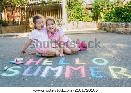 Cheerful children girls sitting near multicolored inscription Hello summer on pavement on sunny day. Sidewalk chalk games for kids concept.