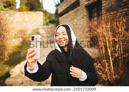 Young happy woman tourist traveling alone and making selfie with attractions by smartphone, have video chat. Blogger broadcasts live stream. Brunette lady in black down jacket and white hoodie. 