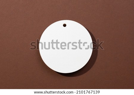 One white colored cardboard brand blank tag of circle shape with little hole in upper part positioned in very center on brown background. Tag mock up. Copy space.