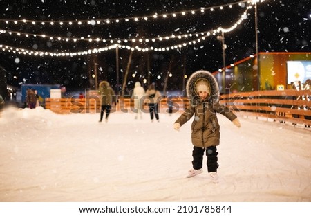 Little girl is skating on ice skates on skating rink in evening, decorated with fairy lights, Christmas trees and fir. Festive mood, Christmas, New Year, holidays, active winter sports and lifestyle