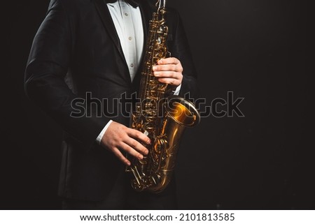 Saxophonist Plays Jazz. Male Musician in a Formal Black Suit Holds a Tenor Saxophone on a Dark Background. Saxophone Close-up. Copy space High quality photo Royalty-Free Stock Photo #2101813585