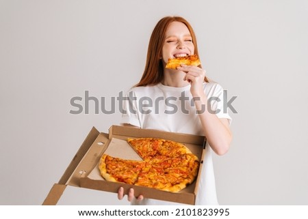 Studio portrait of laughing young woman closed eyes with appetite to eat delicious pizza holding box in hands standing on white isolated background. Pretty redhead female eating tasty meal.
