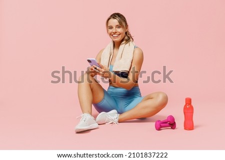 Full body young sporty fitness trainer instructor woman wear blue tracksuit spend time in home gym sit on floor use mobile cell phone isolated on plain light pink background. Workout sport concept. Royalty-Free Stock Photo #2101837222
