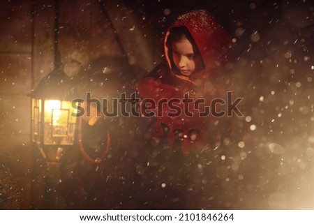 Winter's tale. A little girl in medieval clothes stands on a winter night under a snowfall curiously looking at the burning lantern. Christmas and New Year concept.