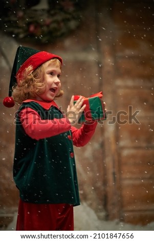 Christmas tale. A portrait of a little elf girl with cute curly hair, who stands by a wooden hut under a snowfall, holds a gift in her hands and laughs. 