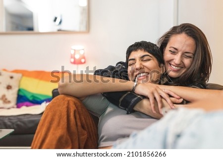 Authentic shot of happy married homosexual female gay couple laughing and embracing on the sofa with rainbow pride flag on background - lesbian couple at home enjoying life together Royalty-Free Stock Photo #2101856266