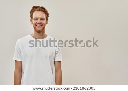 Picture of a smiling young redhead guy with a beard standing over the grey background, hair slicked back, hands down. He wears white T-shirt 