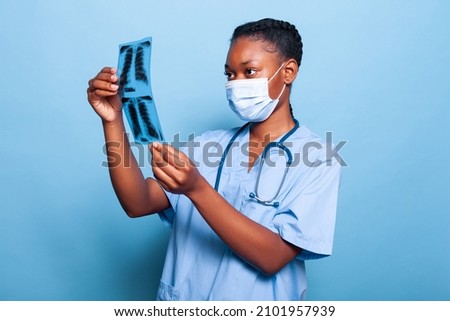 African american practitioner assistant with protection medical face mask to prevent infection with coronavirus analyzing lungs radiography in studio with blue background. Healthcare concept