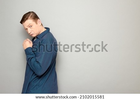 Studio portrait of blond mature man looking shy and timid, feeling guilty, apologizing, asking for forgiveness, wearing casual blue shirt, standing half-turned over gray background, with copy space