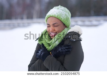 Portrait of frozen suffering girl, young black African Afro American freezing woman standing walking outdoors at winter snowy cold frosty day, shaking, trembling, shivering in jacket, hat, scarf Royalty-Free Stock Photo #2102054875