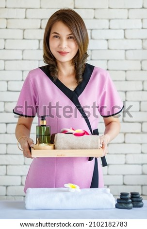 Portrait studio shot of Thai professional female massaging masseuse in uniform stand smiling look at camera hold massage cosmetic perfume oil towel and orchid flower in wood tray on brick background. Royalty-Free Stock Photo #2102182783