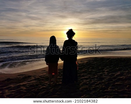 Mother and child stand side by side in silhouette in front of a sunset.