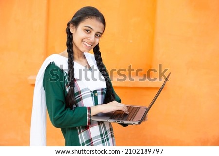 Portrait of Happy young indian school girl using laptop. Smiling braided hair female kid holding computer against orange background, She is looking at camera, skill india concept. Royalty-Free Stock Photo #2102189797