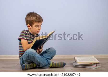 A little boy, studying behind a stack of books and looking at a diary. A male child sitting on the floor on a blue background. Copy space. Royalty-Free Stock Photo #2102207449