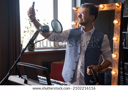 Young smiling singer and pianist is taking a selfie with his phone at a home music studio in front of a piano with a makeup mirror behind his back.