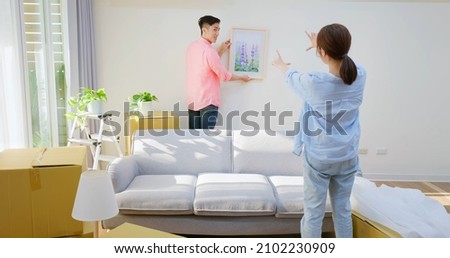 young asian couple decorate home - man hanging photo with frame on wall and woman helping him in living room Royalty-Free Stock Photo #2102230909