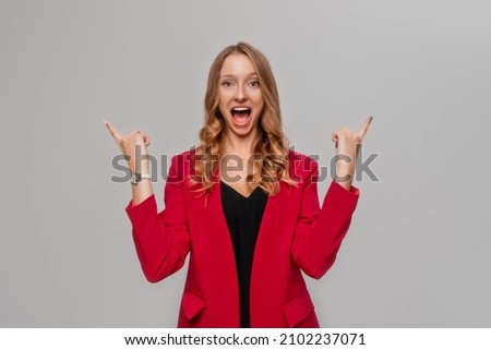 Hey look there. Smiling excited blonde young woman pointing fingers up, showing to advertisement, promo offer, stands in red blazer against gray background